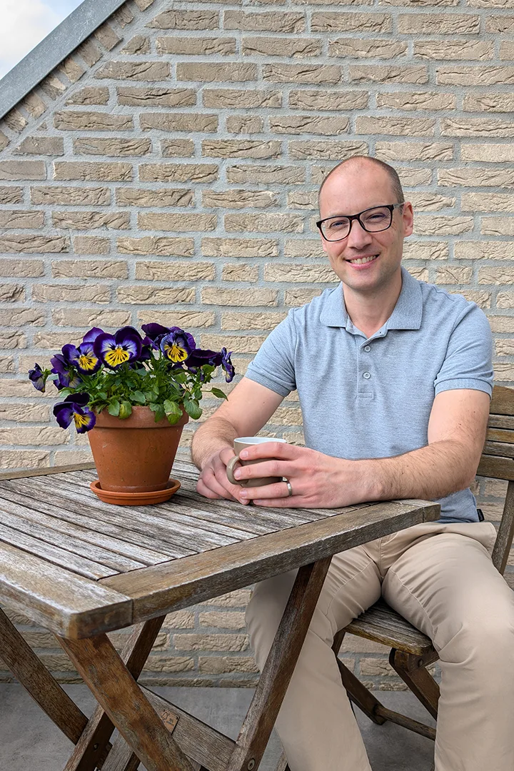 Michiel Kox aan een houten tafel op zijn terras, met een kop koffie